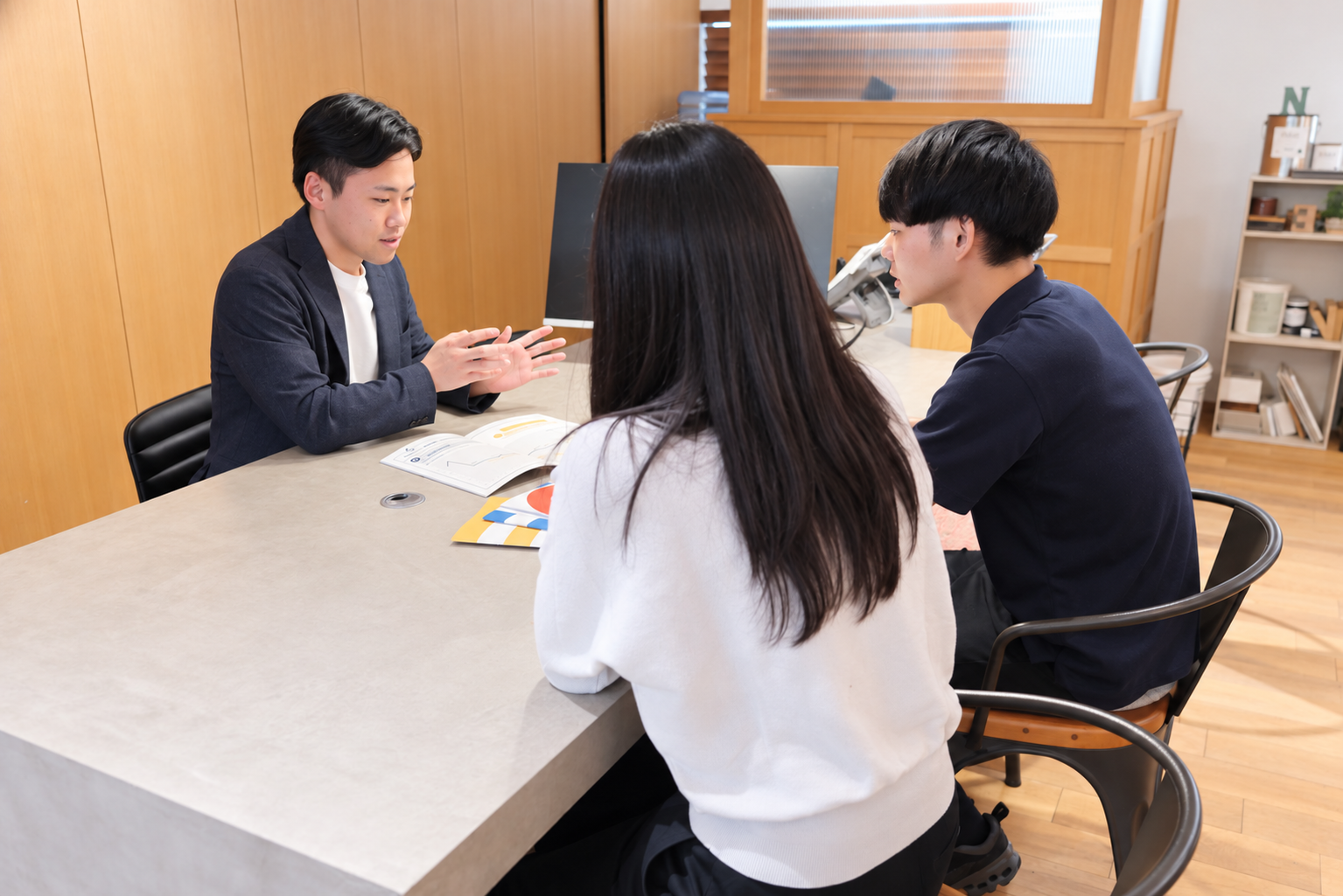 木目の壁の店舗で、テーブル越しにスタッフが資料を使ってカップルに説明している様子